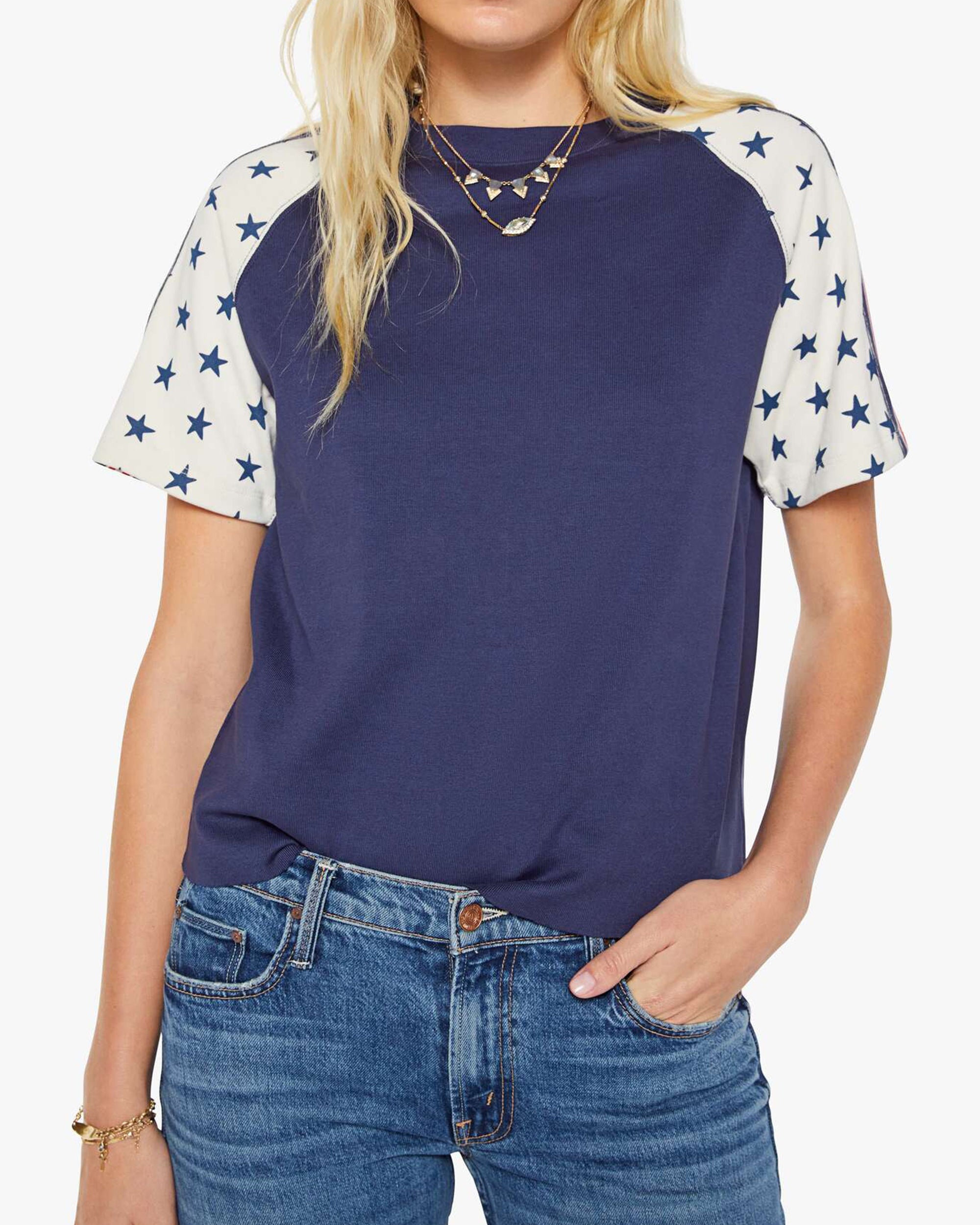 A woman wears The SS Concert Race by Mother, a boxy fit 100% cotton crewneck tee with navy and white star-patterned raglan sleeves, paired with blue jeans and layered necklaces. One hand is in her pocket.