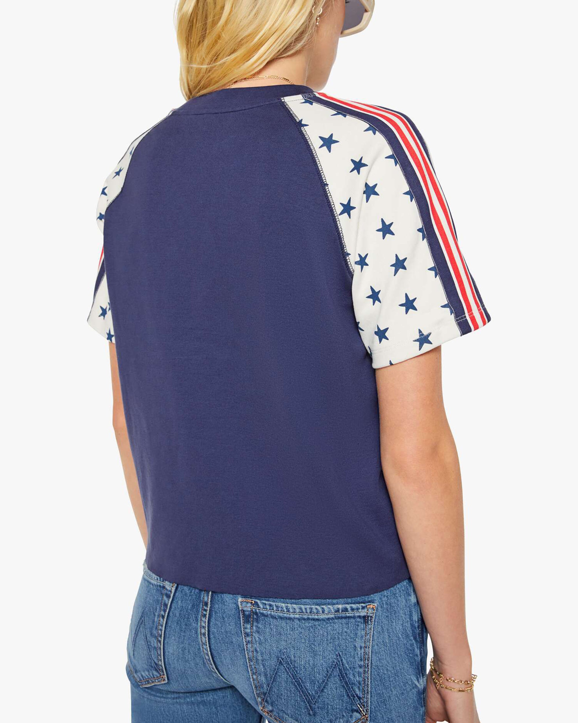 A person wearing Mother’s The SS Concert Race tee—a navy blue raglan top with cream sleeves, blue stars, and red stripes—in 100% cotton with a boxy fit, styled with blue jeans and shown from the back.