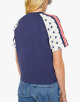 A person wearing Mother’s The SS Concert Race tee—a navy blue raglan top with cream sleeves, blue stars, and red stripes—in 100% cotton with a boxy fit, styled with blue jeans and shown from the back.