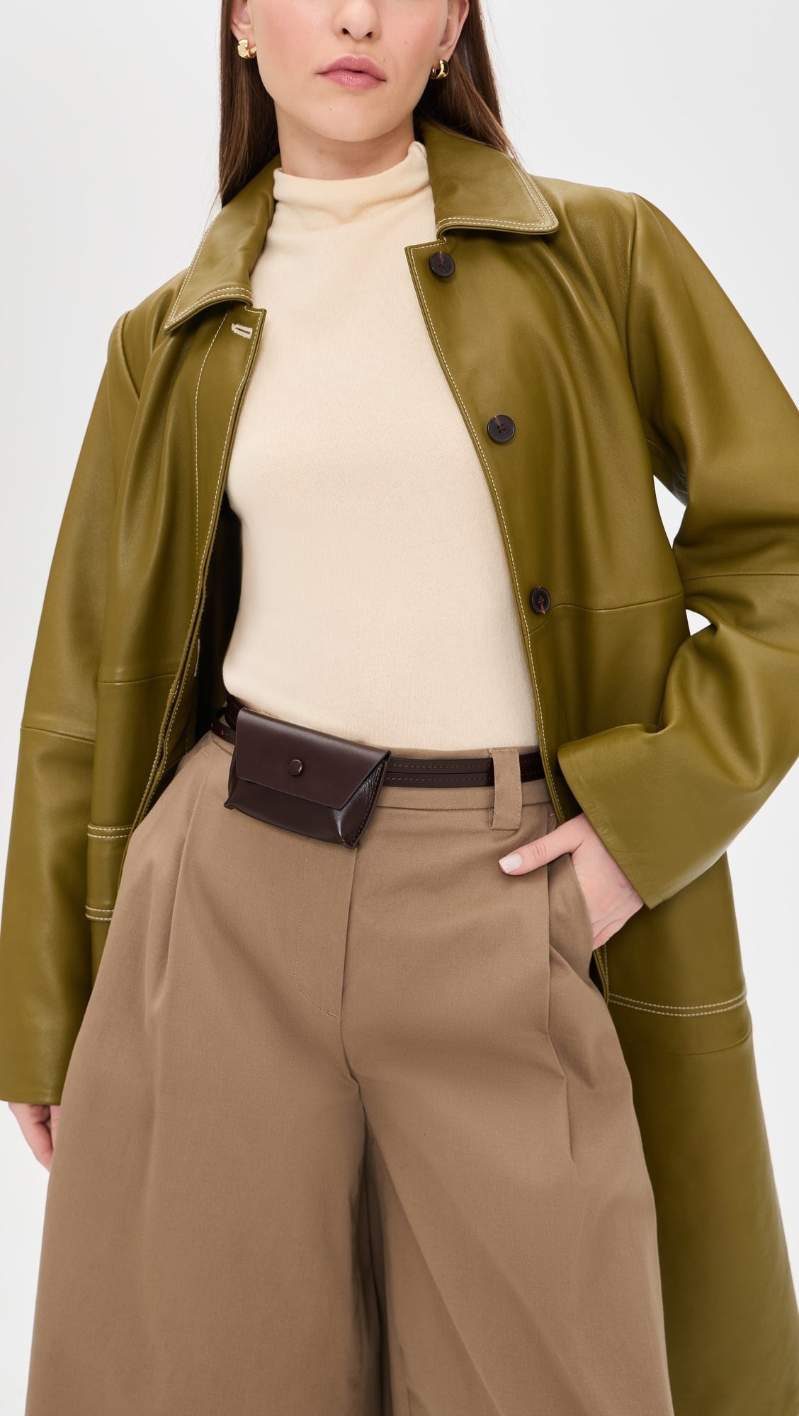 A woman in an olive green leather coat, beige turtleneck, and tan wide-leg pants stands with one hand in her pocket, accessorized with the Rachel Comey Double Wrap Hamcon Belt with Pouch featuring gold-tone hardware.