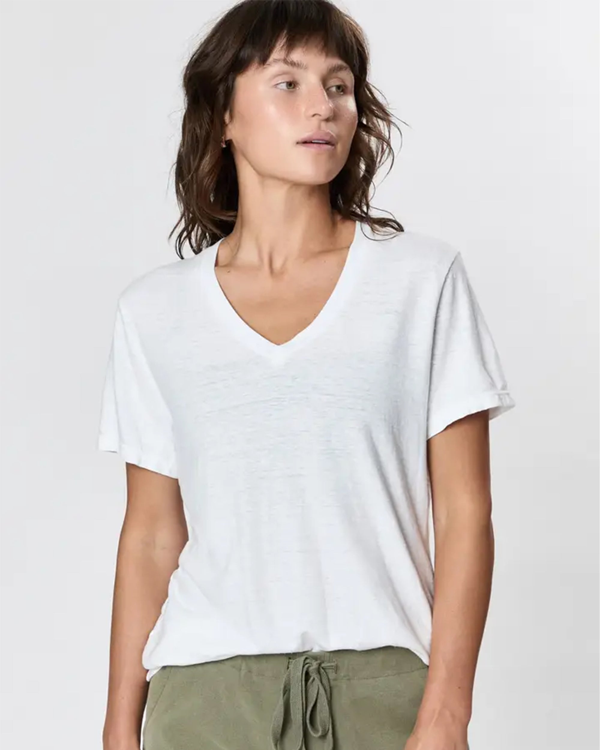 A person with wavy brown hair wears the Faire Hemp V Neck, a loose white T-shirt, and olive green drawstring pants, standing against a plain light background and looking slightly to the side.