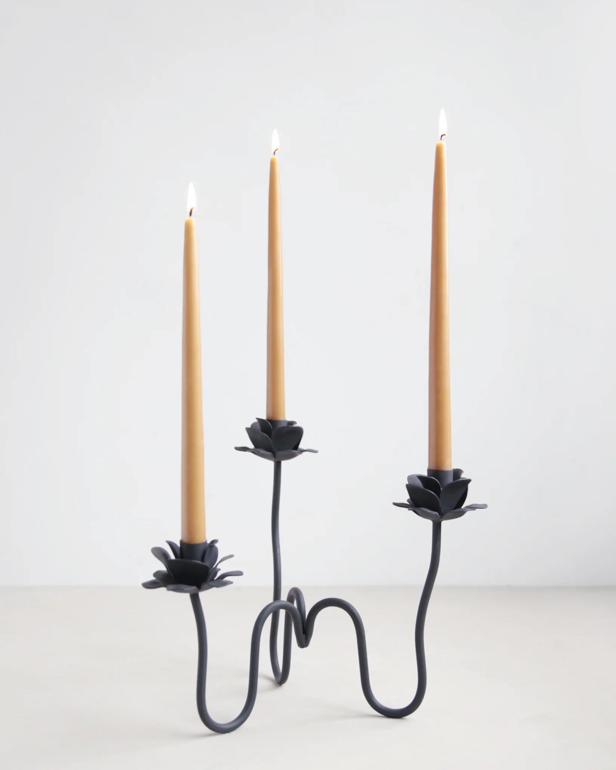 The Floral Society's Rana Candlebra is a black, hand-forged iron candelabra with three arms and flower-like holders, each displaying a tall tan taper candle. Perfect for elegant decor, it stands out against a plain white background.