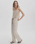 A woman with wavy hair poses casually against a plain background, wearing the Rag & Bone rb miramar trry drwcrd wde leg cream-colored cotton terry pants with a drawstring waist, a beige sleeveless top, and black sandals.