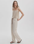 A woman with wavy hair poses casually against a plain background, wearing the Rag & Bone rb miramar trry drwcrd wde leg cream-colored cotton terry pants with a drawstring waist, a beige sleeveless top, and black sandals.