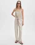 A woman in Rag & Bone rb miramar trry drwcrd wde leg pants, sunglasses, a beige spaghetti-strap top, and black flip-flops stands against a plain white background.