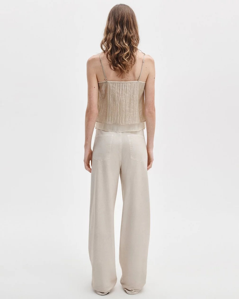 A woman with wavy, shoulder-length hair faces away, modeling Rag &amp; Bone’s rb miramar trry drwcrd wde leg wide-leg jeans with a pull-on drawstring waistband and a beige spaghetti strap fitted top, against a plain white background.