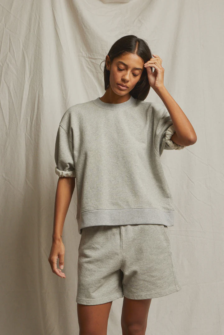 A woman stands before a beige fabric backdrop in the Perfectwhitetee Rebecca SS Crew French terry cotton sweatshirt and matching shorts, her straight dark hair falling as she touches her head and looks down.
