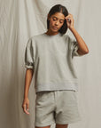 A woman stands before a beige fabric backdrop in the Perfectwhitetee Rebecca SS Crew French terry cotton sweatshirt and matching shorts, her straight dark hair falling as she touches her head and looks down.