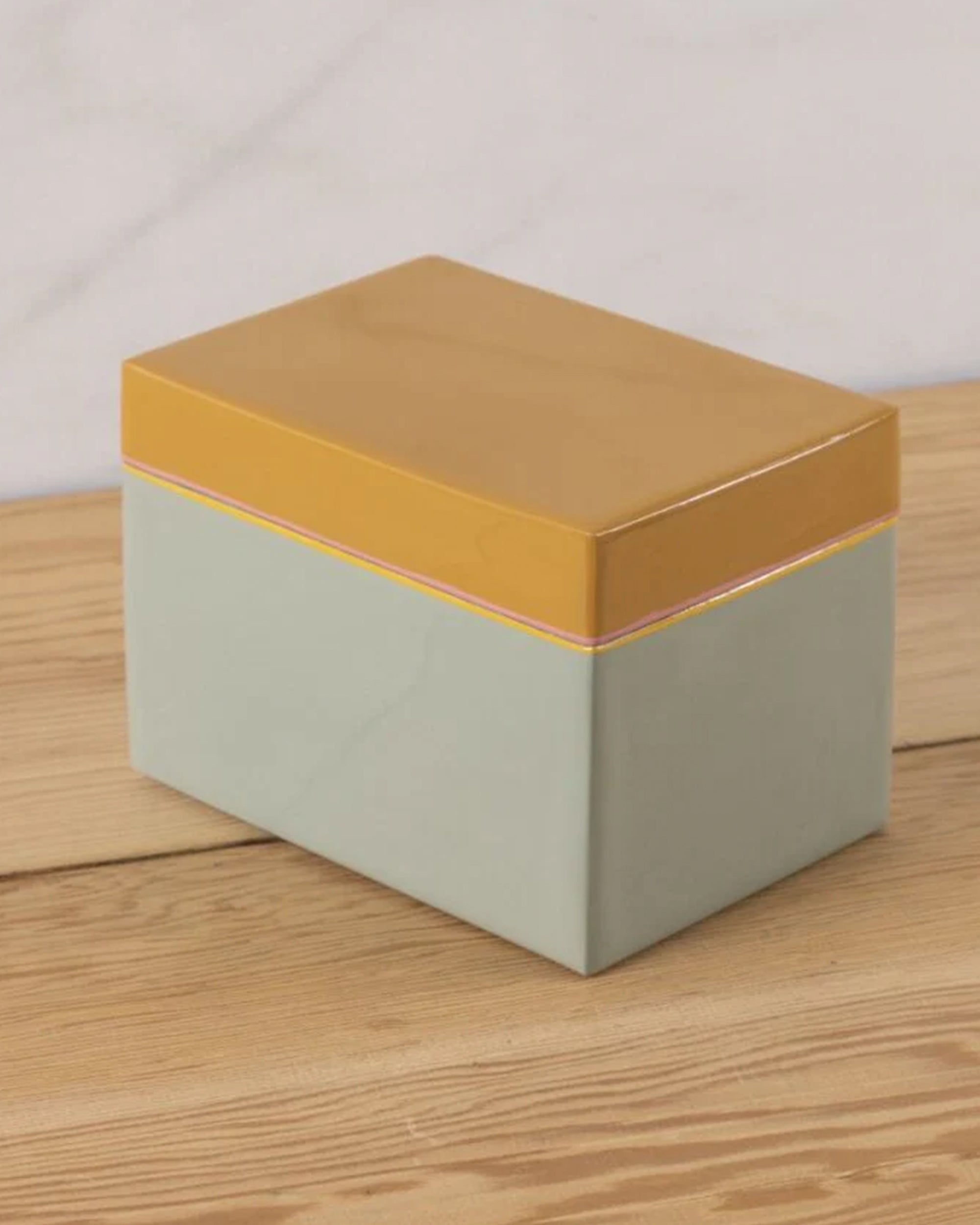 The Harlow Bright Recipe Box by Be Home, featuring a mustard yellow lid and pale green base, sits on a wooden surface and brings gourmet kitchen charm against a light background.