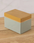 The Harlow Bright Recipe Box by Be Home, featuring a mustard yellow lid and pale green base, sits on a wooden surface and brings gourmet kitchen charm against a light background.