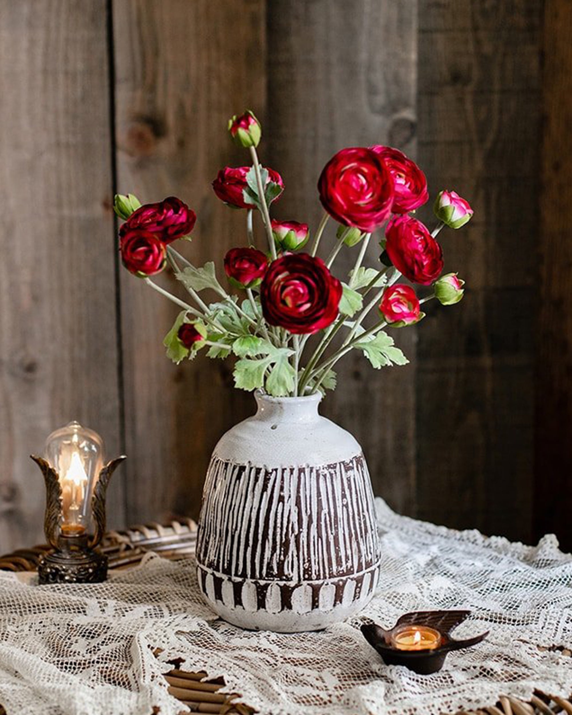 A white vase with dark brown vertical stripes holds Faire’s Buttercup Flower Bundle Red Burgundy on a lace-covered table. Nearby are a lit candle in a small holder and a vintage-style bulb, all set against a rustic wooden background.