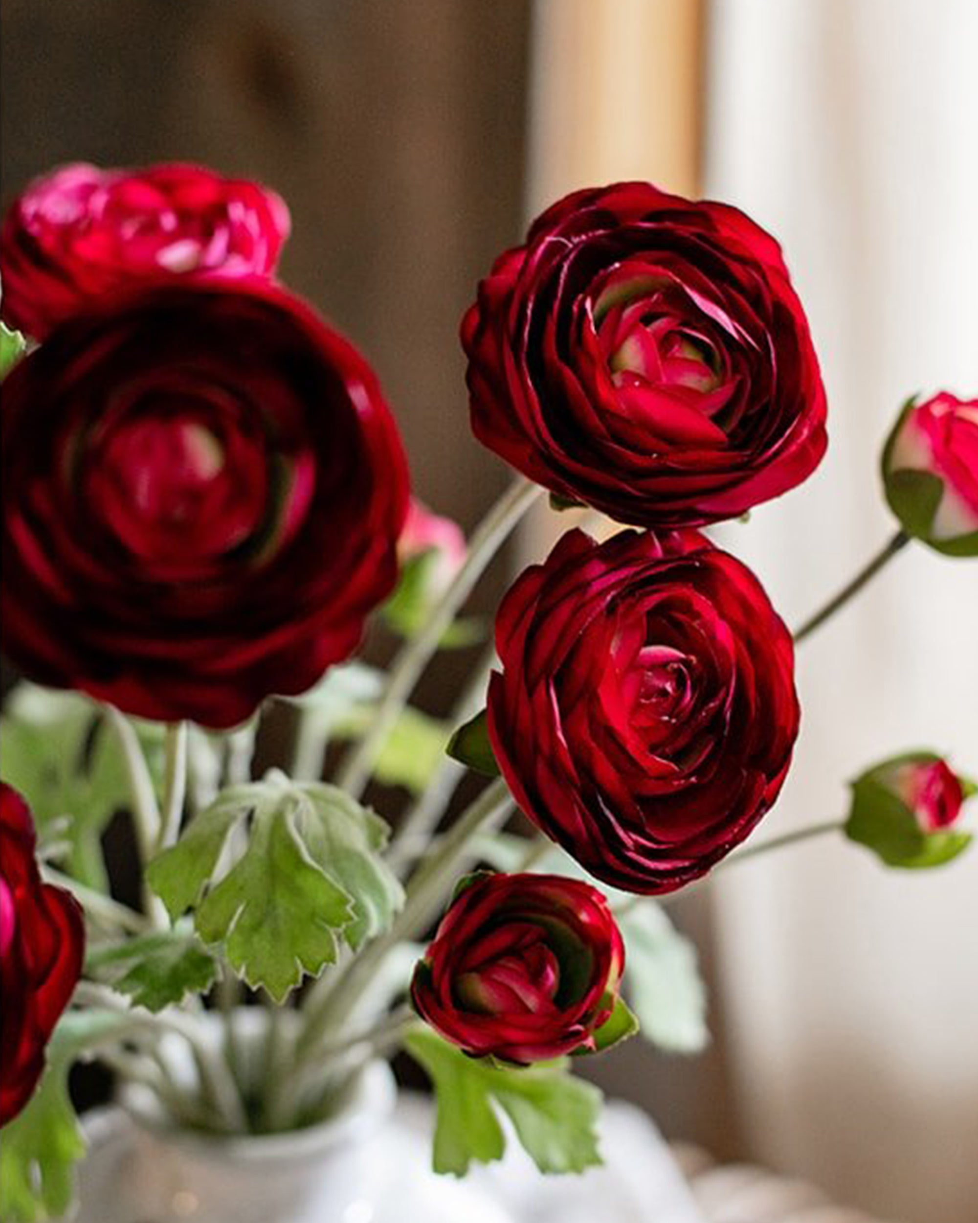 The Buttercup Flower Bundle Red Burgundy by Faire features deep red faux silk flowers with green leaves in a white vase, gently illuminated by natural window light.
