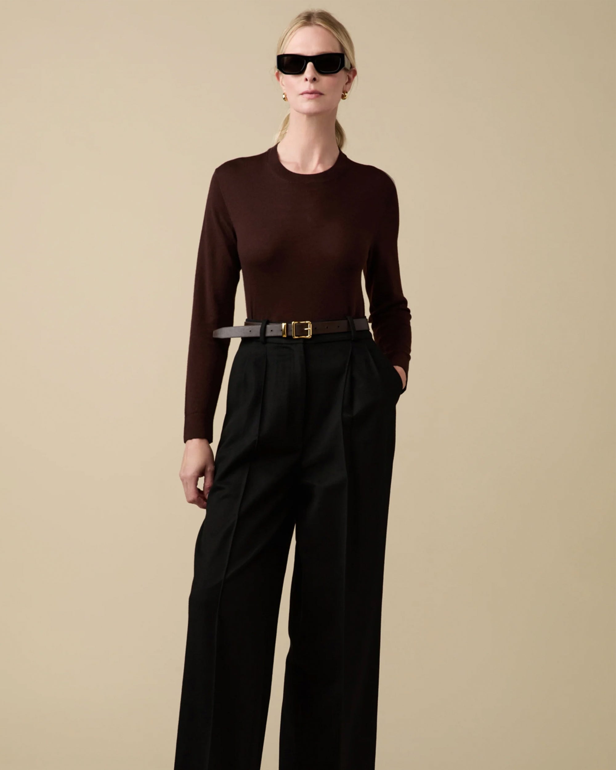 Wearing black sunglasses and the Sold Out NYC Refined Crew eco-friendly dark brown Merino wool sweater, tucked into high-waisted black wide-leg trousers with a black belt, against a beige background.