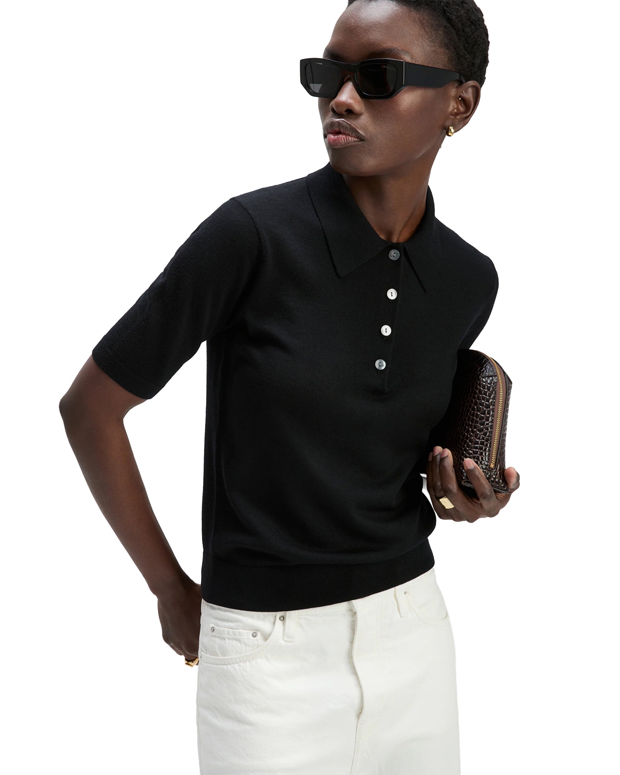 Wearing sunglasses, The Refined SS Polo by Sold Out NYC in black with buttons, and white pants, a short-haired person with gold earrings holds a textured brown clutch.