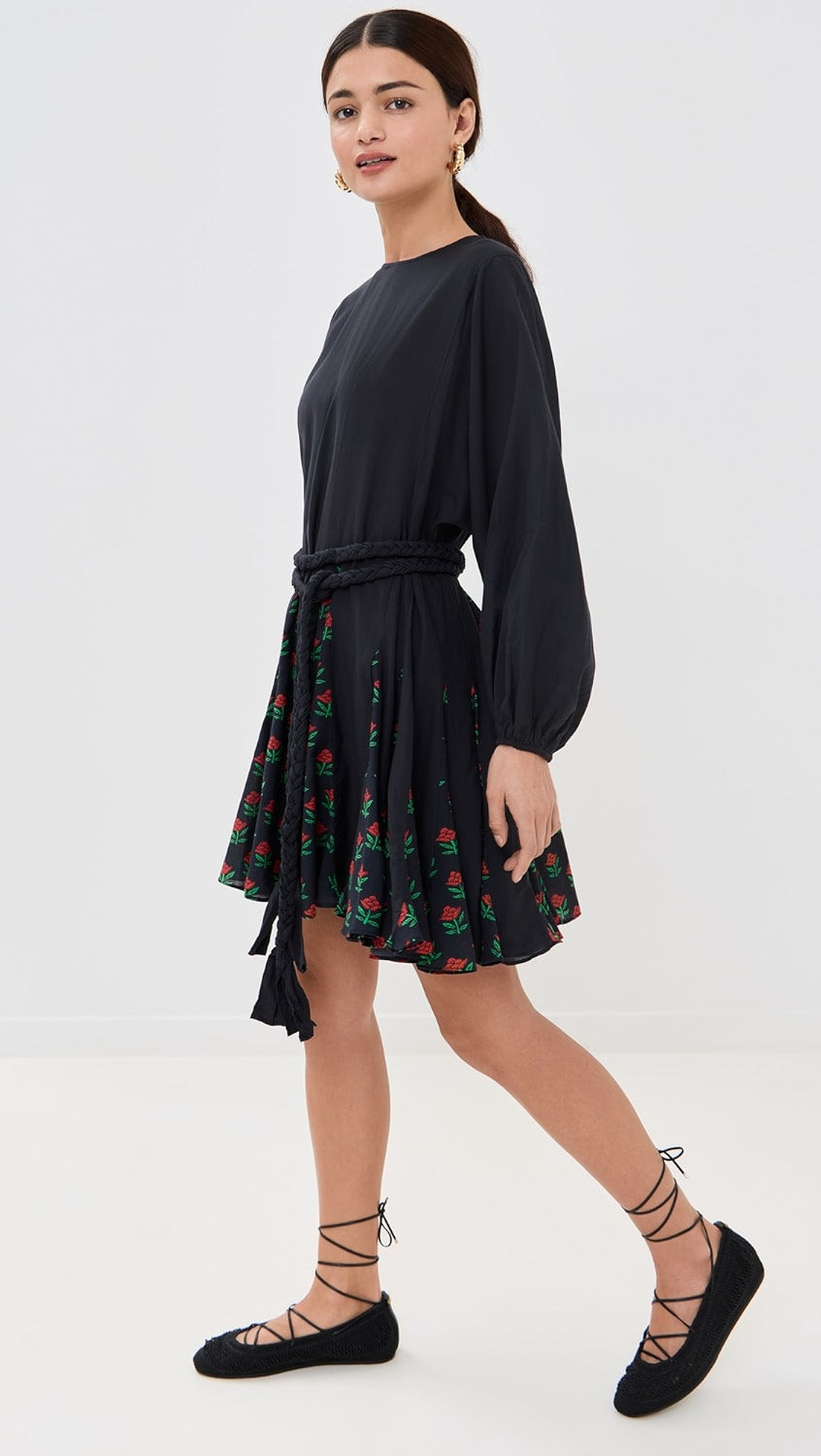 A person is posing in front of a plain backdrop wearing the Ella Dress by Rhode, featuring floral printed panels and a braided belt. They showcase the dress's lightweight weave while looking slightly to the side, paired with black lace-up shoes.