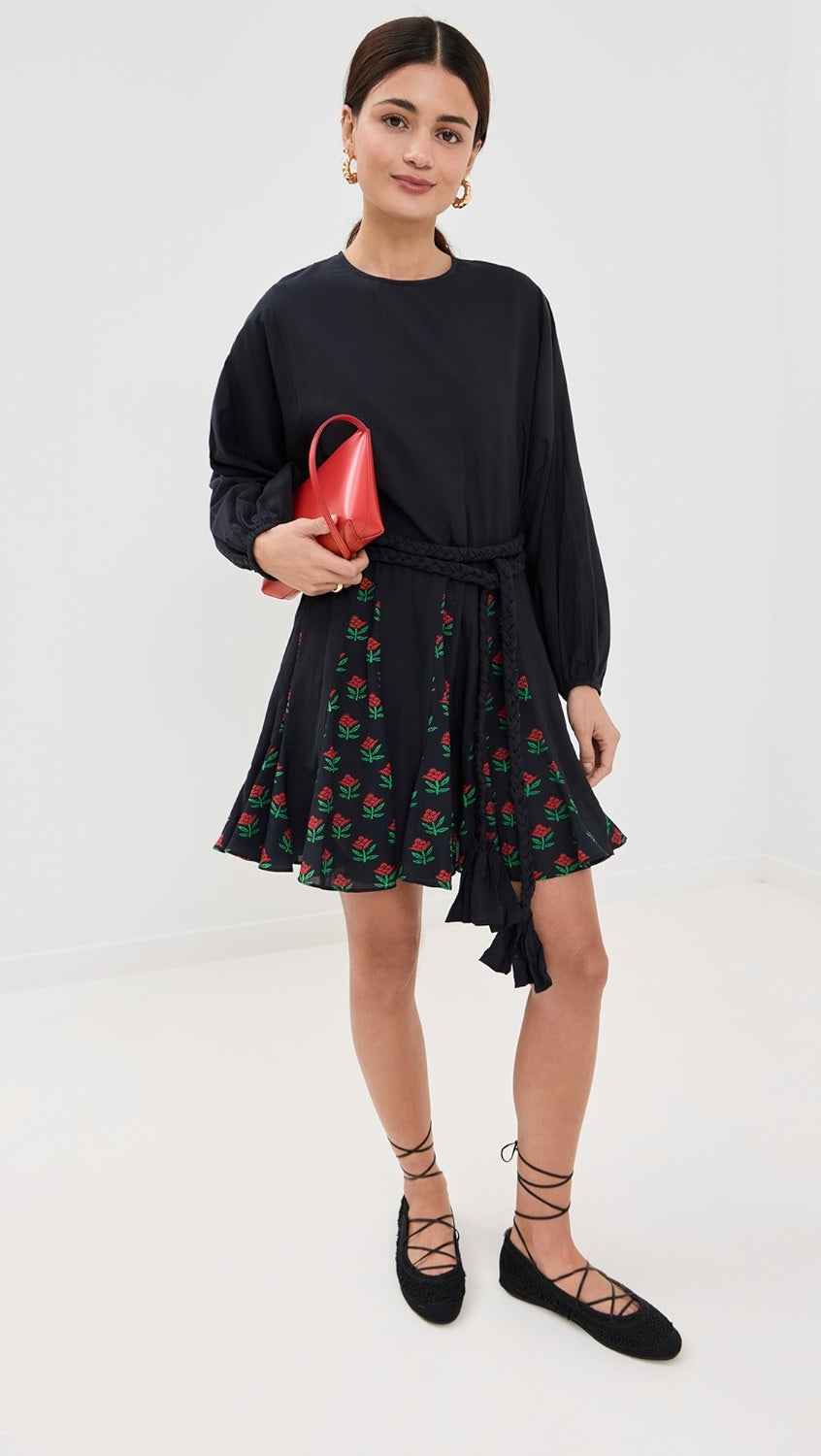 In a white room, a person wears the Rhode Ella Dress, a black long-sleeve top, floral skirt panels, and black lace-up shoes. They're accessorized with gold hoop earrings and hold a small red handbag while their dark hair is styled in braided ties.