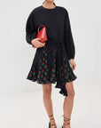 In a white room, a person wears the Rhode Ella Dress, a black long-sleeve top, floral skirt panels, and black lace-up shoes. They're accessorized with gold hoop earrings and hold a small red handbag while their dark hair is styled in braided ties.