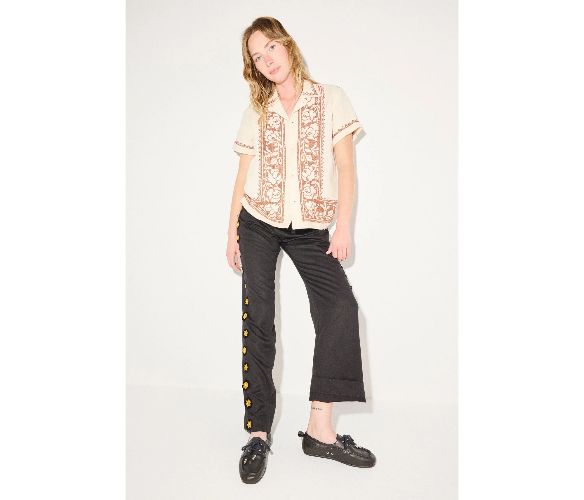 A woman with light brown hair poses casually against a white background, wearing a cream embroidered short-sleeve shirt, black pants with gold buttons, and Freda Salvador’s RIA Driver Loafers in stylish black Nappa leather.
