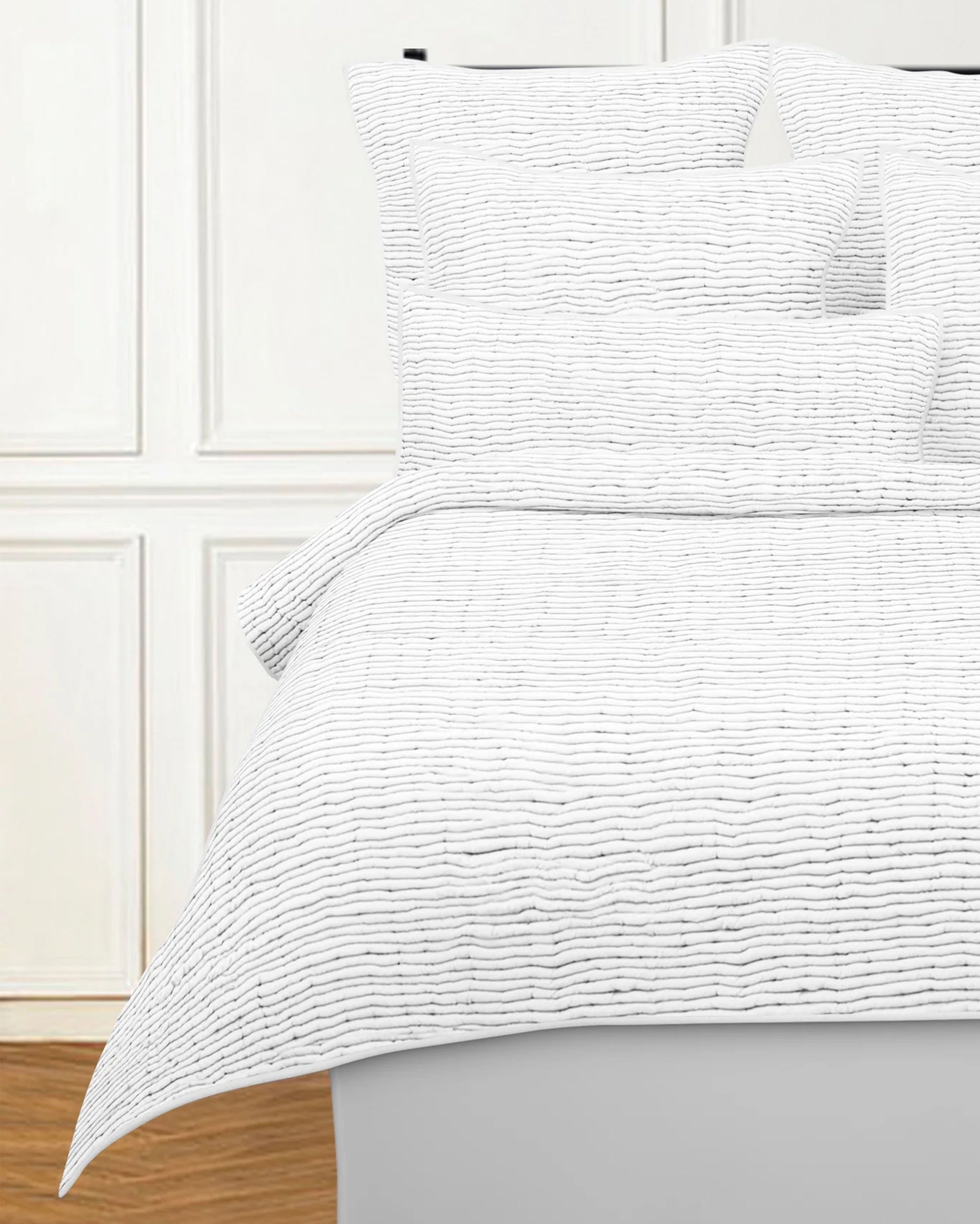 A neatly made bed features the Avasa Bedding Ridge Quilt White King and matching shams, set against a white paneled wall. The wooden floor and bright, minimalistic room create a cozy, inviting atmosphere.