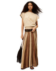 A woman with long curly hair wears the BA&SH Rima Skirt and a beige top, paired with brown sandals, a black belt, and a fringed black bag. She accessorizes with a necklace and bracelets.