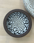 The Ritual Bowls 4.5" by Faire feature a round ceramic design with a dark brown rim and white interior accented by an irregular black-outlined crackle pattern—perfect as versatile bowls or chic salt cellars on light wood surfaces.