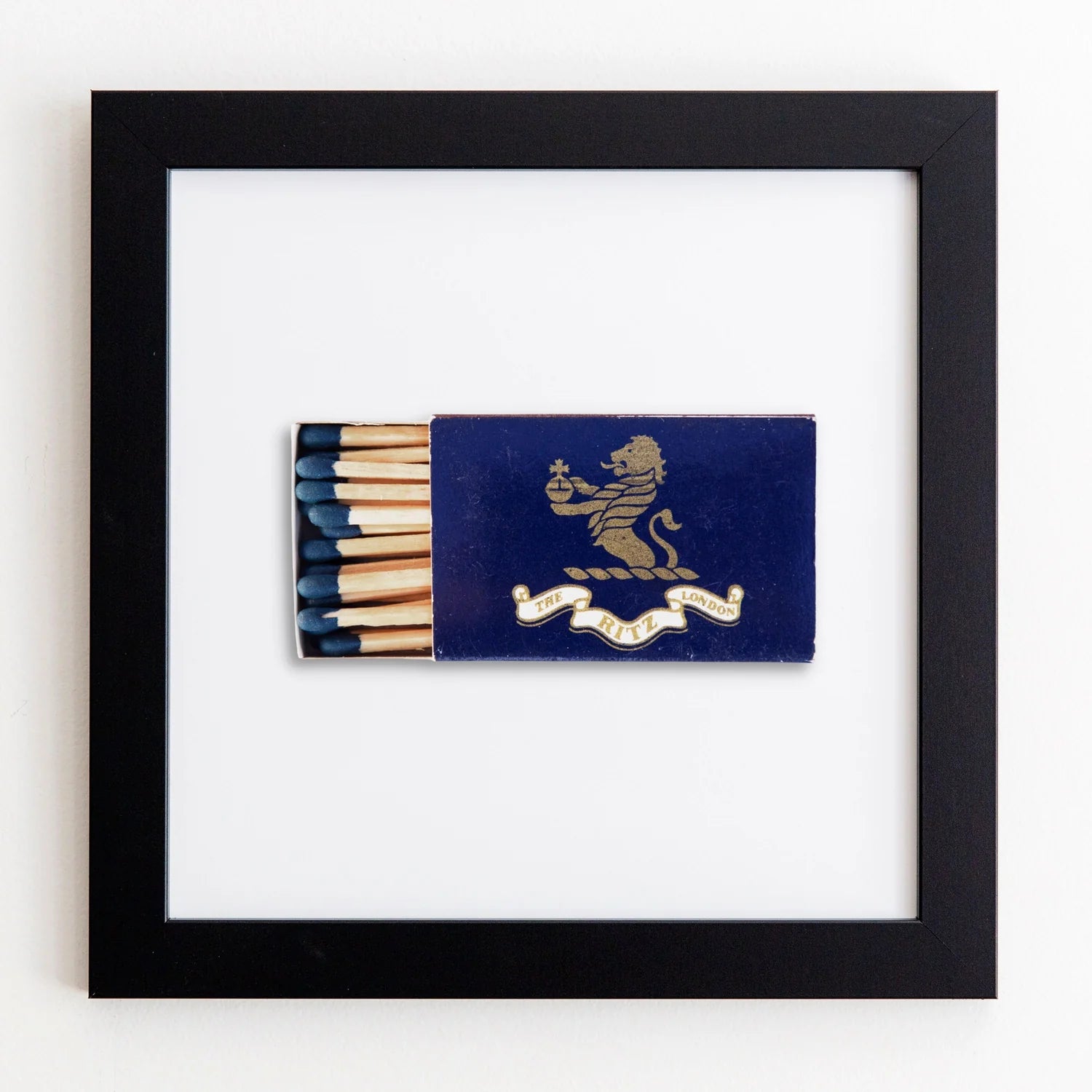 A framed artwork featuring a blue Match South matchbook with a gold lion emblem, open to display several matches, against a white acrylic background in an Art Square Black Frame.