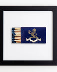 A framed artwork featuring a blue Match South matchbook with a gold lion emblem, open to display several matches, against a white acrylic background in an Art Square Black Frame.