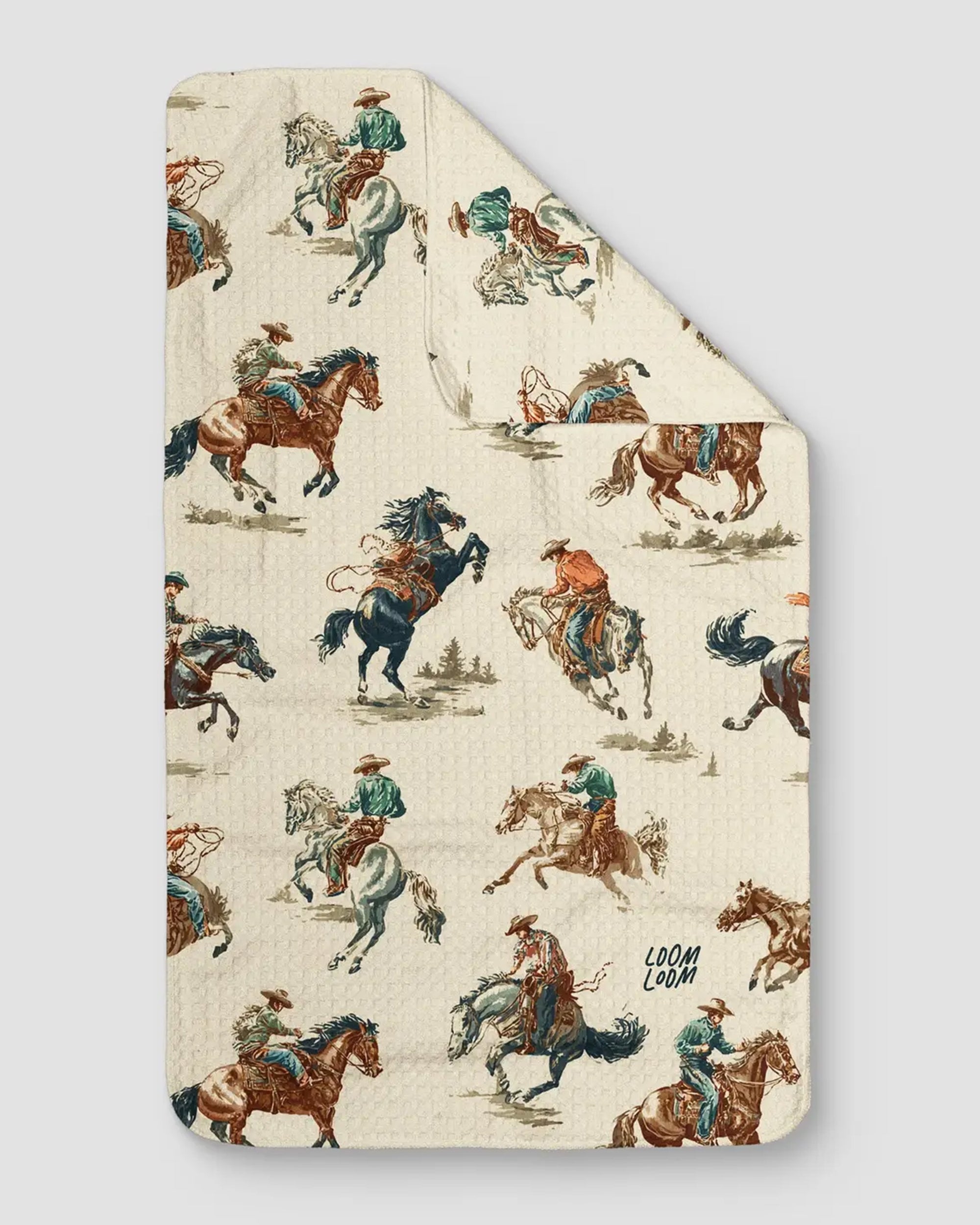The Rodeo Towel by Faire is a beige towel made from EcoLoom fabric, showcasing a vintage western pattern of cowboys riding and roping horses in muted colors.