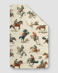 The Rodeo Towel by Faire is a beige towel made from EcoLoom fabric, showcasing a vintage western pattern of cowboys riding and roping horses in muted colors.