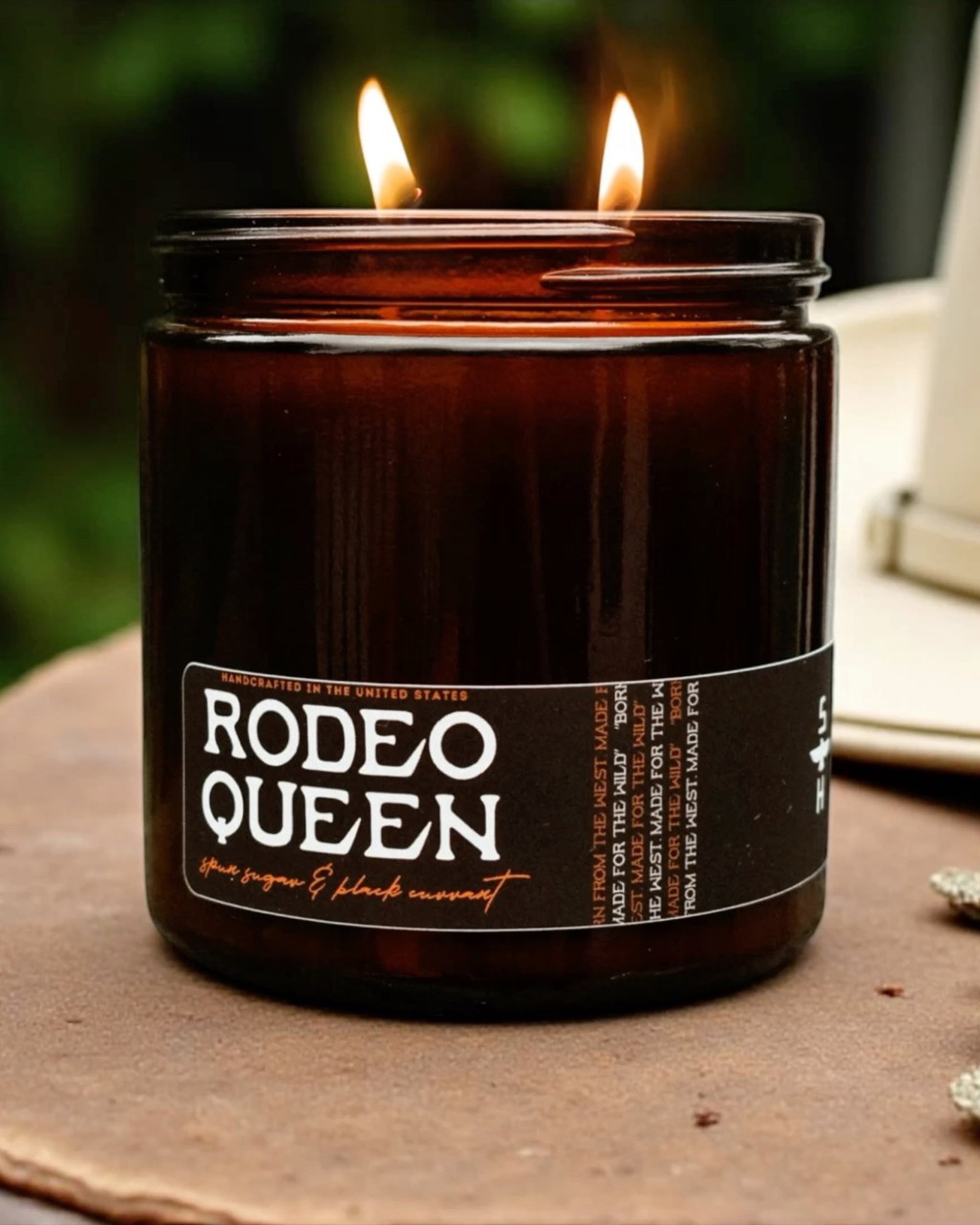 A brown glass Faire candle labeled "Black Currant & Spun Sugar" with two flames burns outdoors. A white object, possibly a hat, is partially visible in the background.