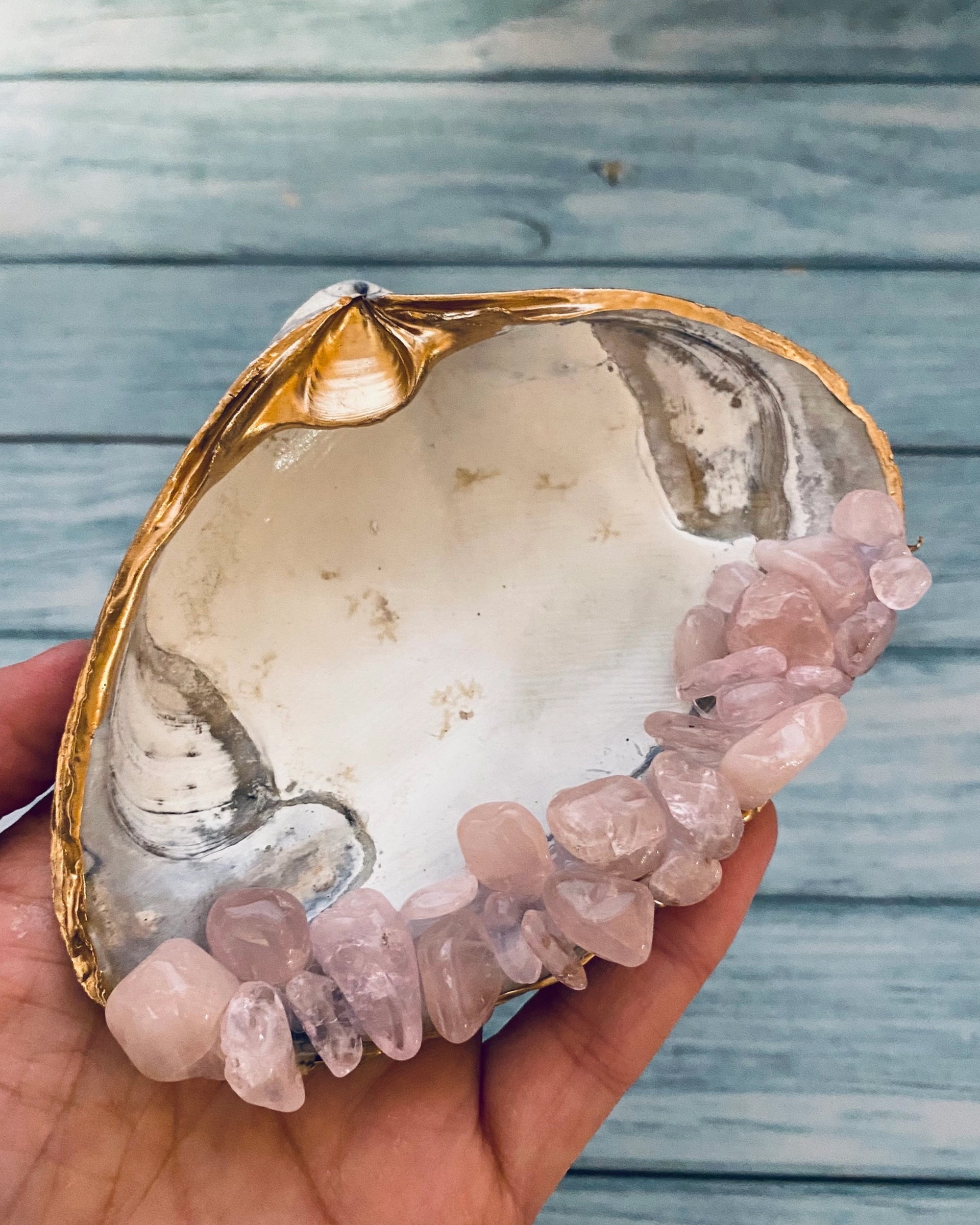 A hand holds the Faire Rose Quartz Shell, a large handmade shell with a gold-edged rim and polished light pink crystals, set against a blue wooden background.