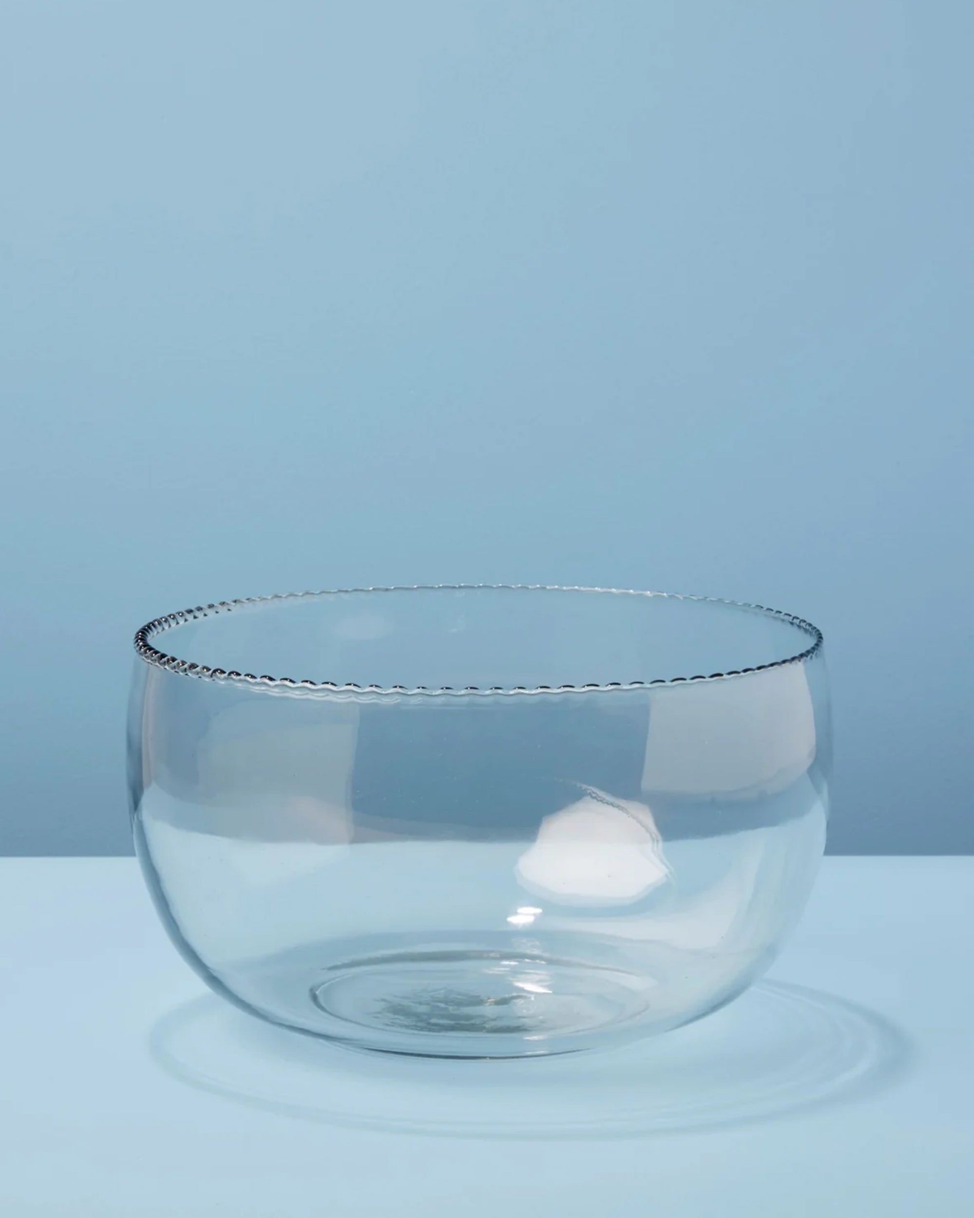The Be Home Ruffle Glass Bowl L, featuring a clear beaded rim inspired by ruffle glassware, rests on a light blue surface against a blue background.