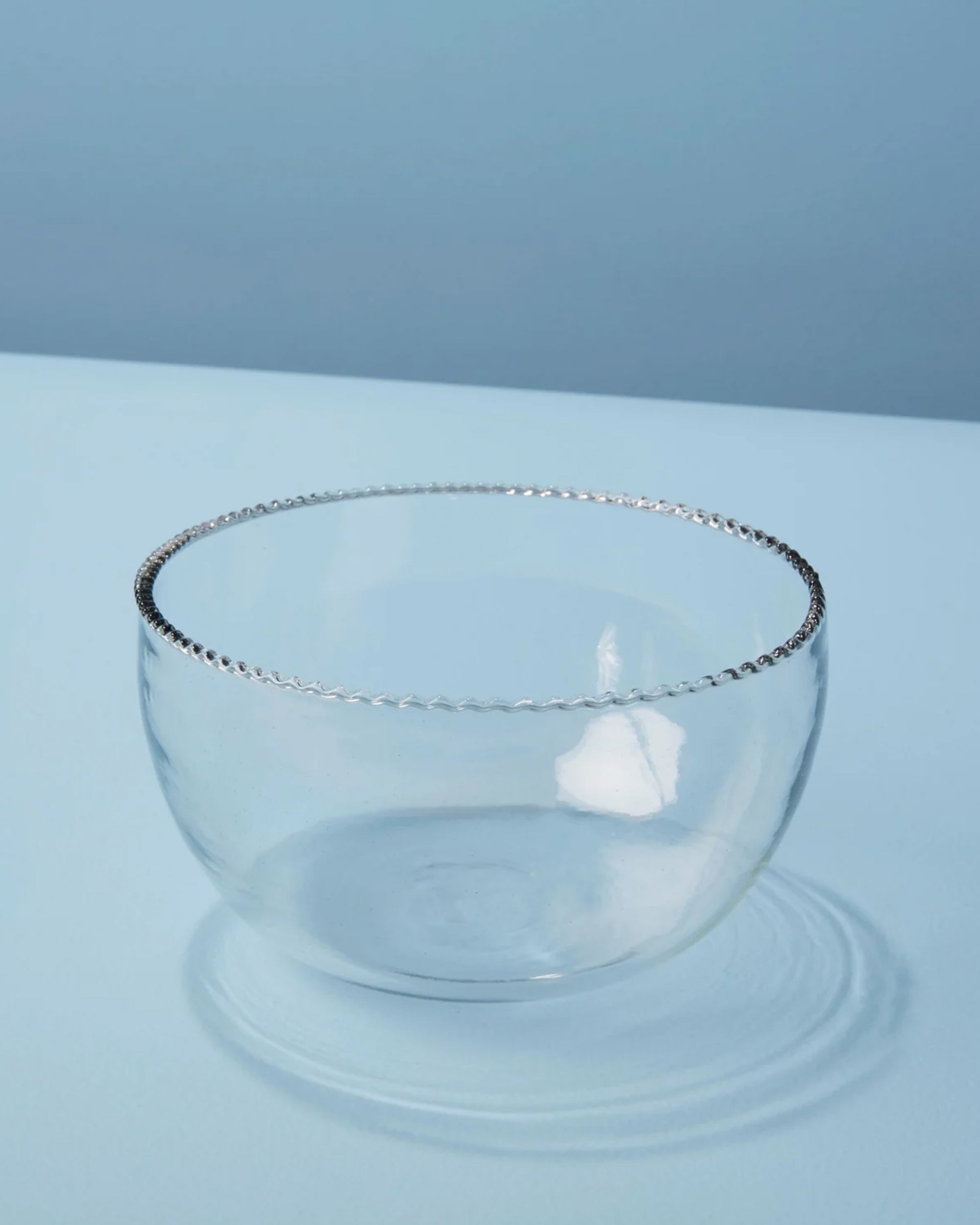 The Be Home Ruffle Glass Bowl S, with its elegant beaded rim, is displayed on a smooth light blue surface against a matching background, highlighting the beauty of this clear glassware piece.