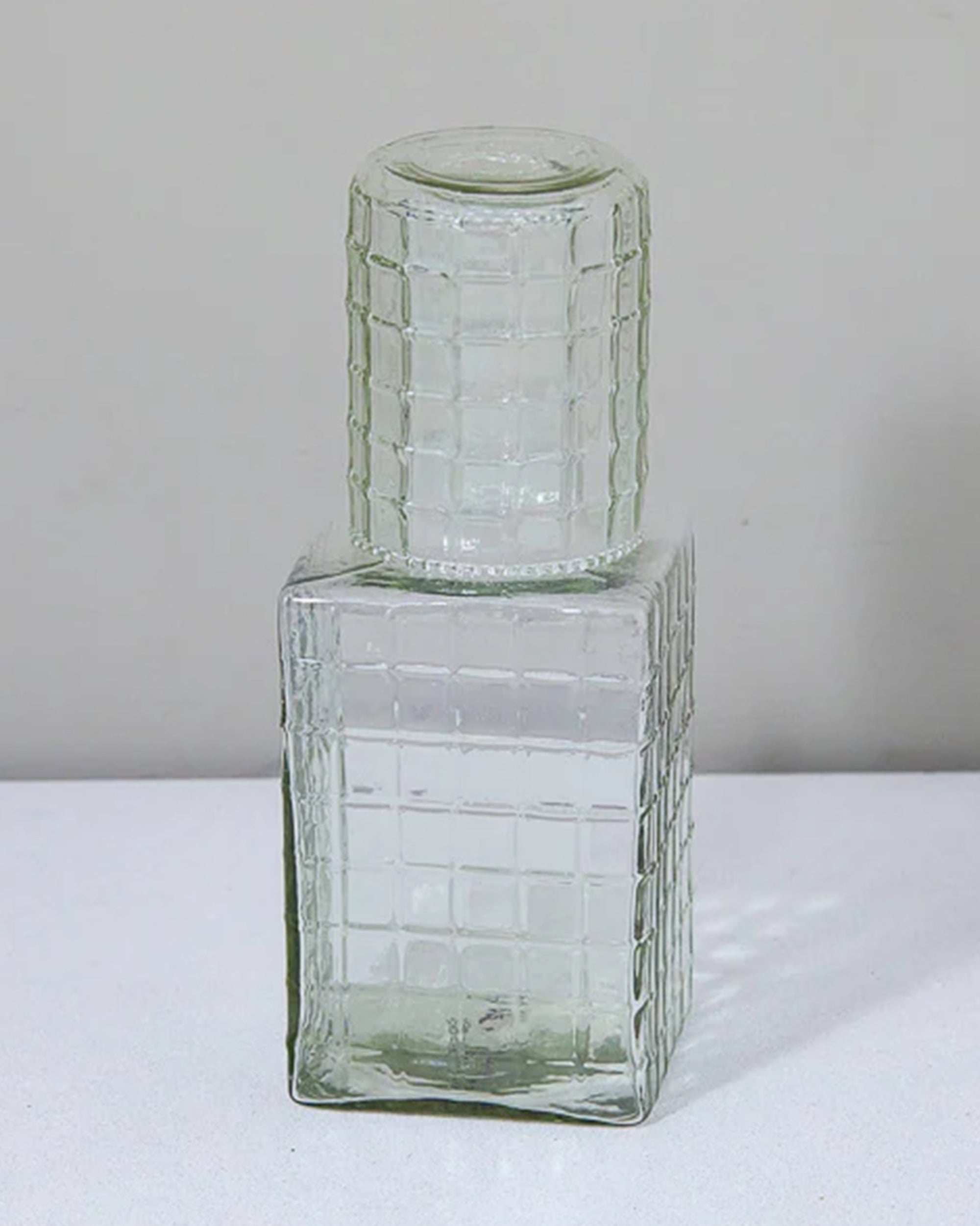 The Be Home Ruffle Glass Bedside Set includes a clear, square carafe with a matching lid, both with a grid pattern. Stylish and chic, it's the perfect nightstand accessory, shown here on a white surface against a light gray background.