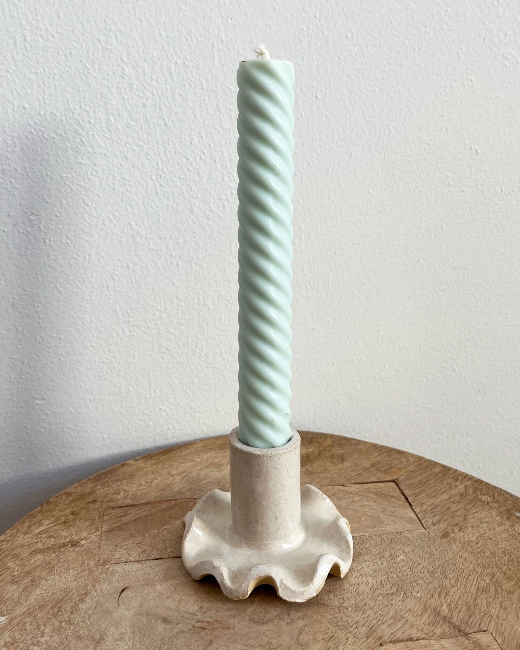 The Faire Ruffle Candle Holder, an off-white ceramic piece with wavy edges, holds a light blue twisted candle upright on a wooden surface against a plain white wall.