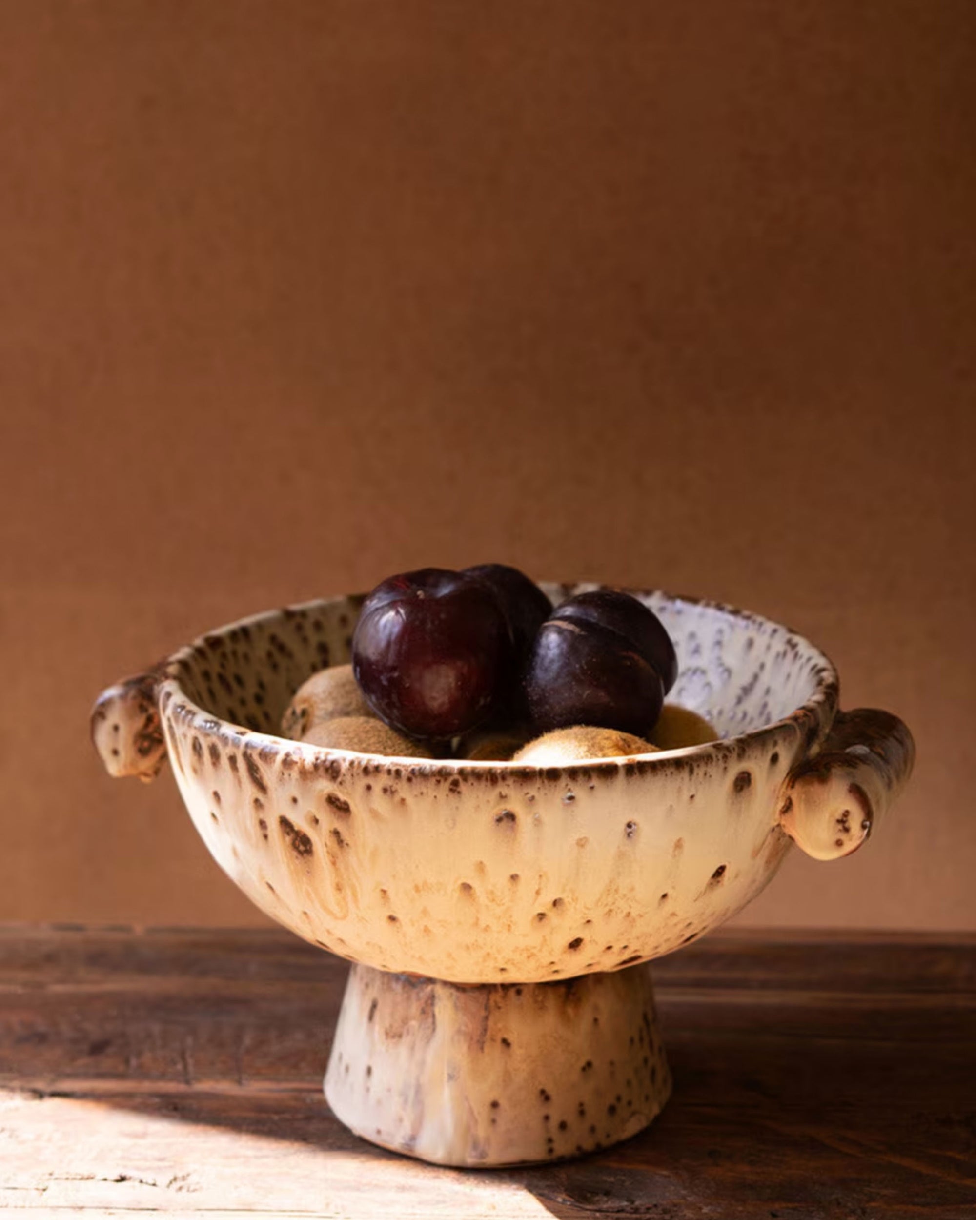 The CFNG1002 Rustic Ceramic Compote by Kalalou, Inc features two handles and holds dark plums and light fruits on a wooden surface—ideal kitchen decor against a plain brown background.