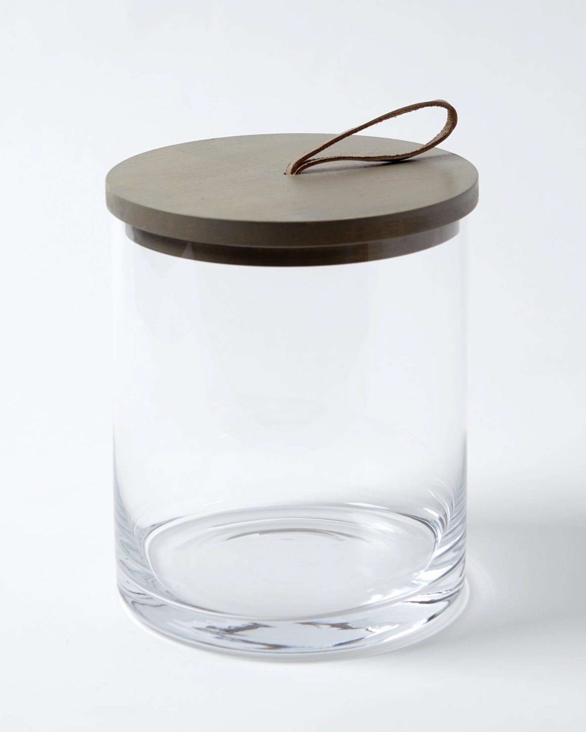 The BB-6.60026 Rustic Canister MD by Global Views features clear glass, a round flat beige lid, and a looped brown leather handle—ideal for modern or rustic kitchen storage—displayed against a white background.