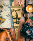 The Faire Saguaro Ntnl Park Pennant, featuring a vintage felt design with a saguaro cactus and "EST 1994," rests on a wooden table with a map, camera, compass, coffee cup, and leather bag.