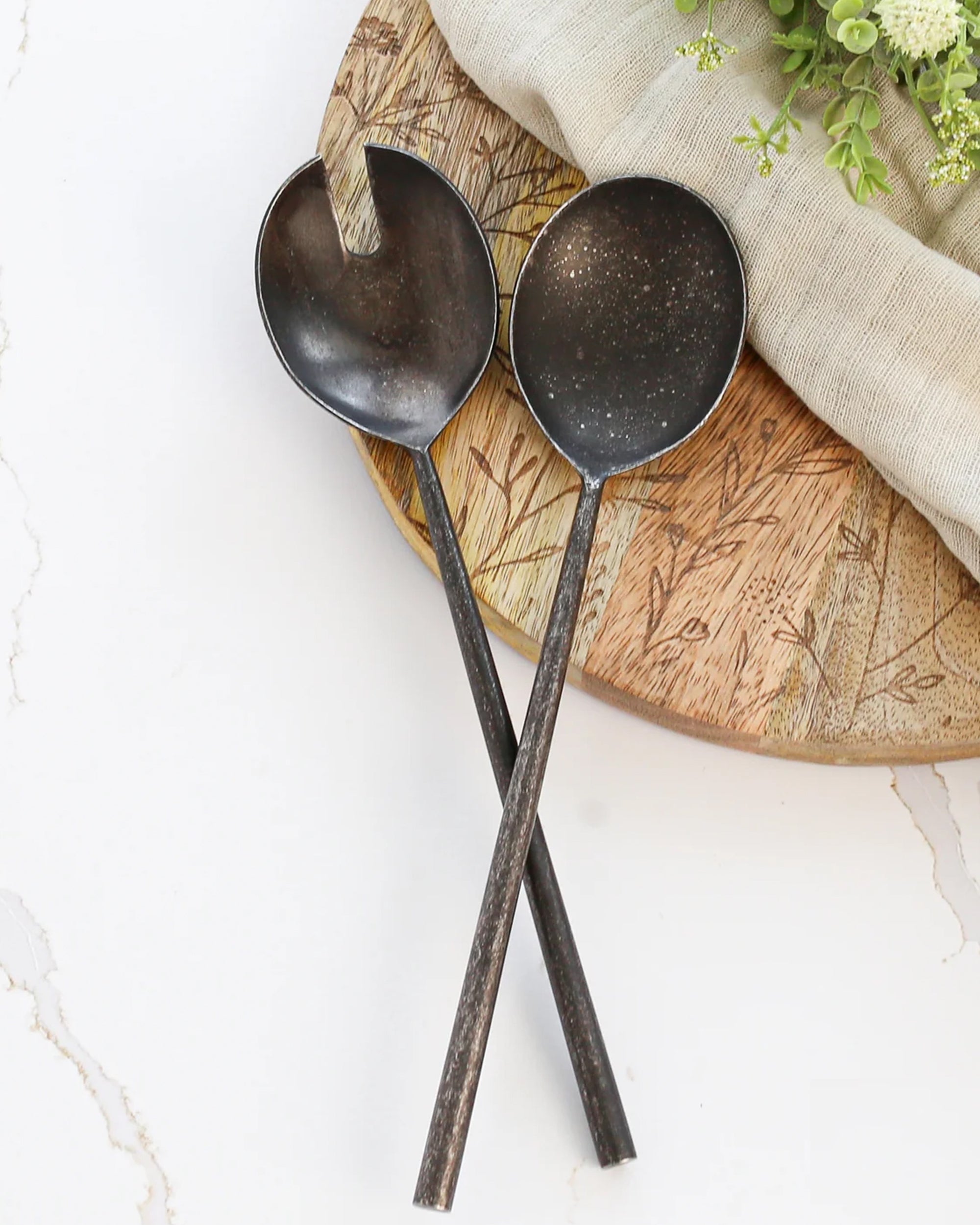The Indaba 4-7231 Tides Salad Servers S/2 Black, featuring hand-forged dark metal and long handles, are crossed on a round wooden board beside beige cloth and green foliage, all displayed on a white marble surface.