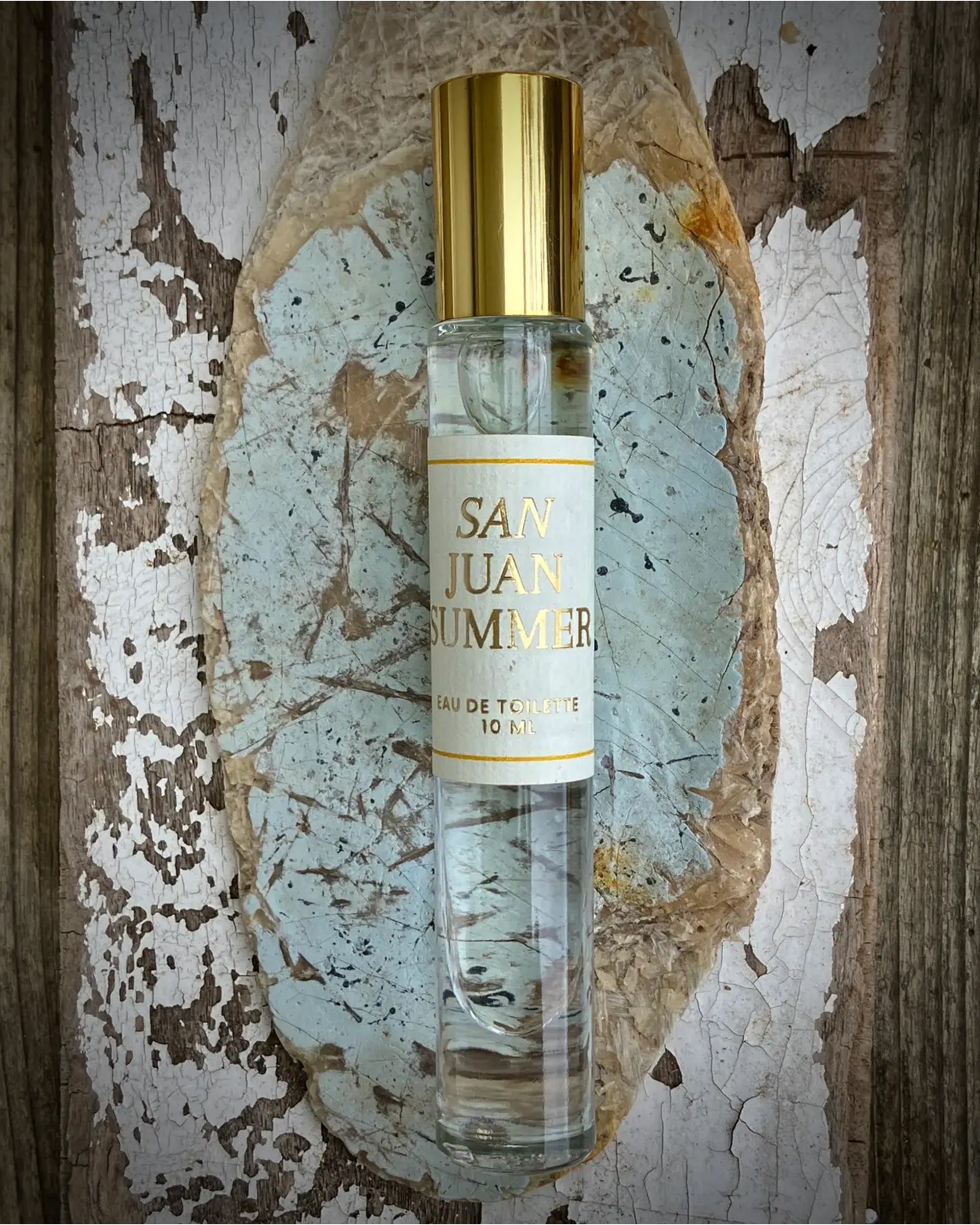 A clear glass perfume bottle labeled "San Juan Roll On" by Faire with a gold cap sits on a cracked blue and white painted stone, framed by weathered wood—evoking the scent of a citrus-kissed breeze.