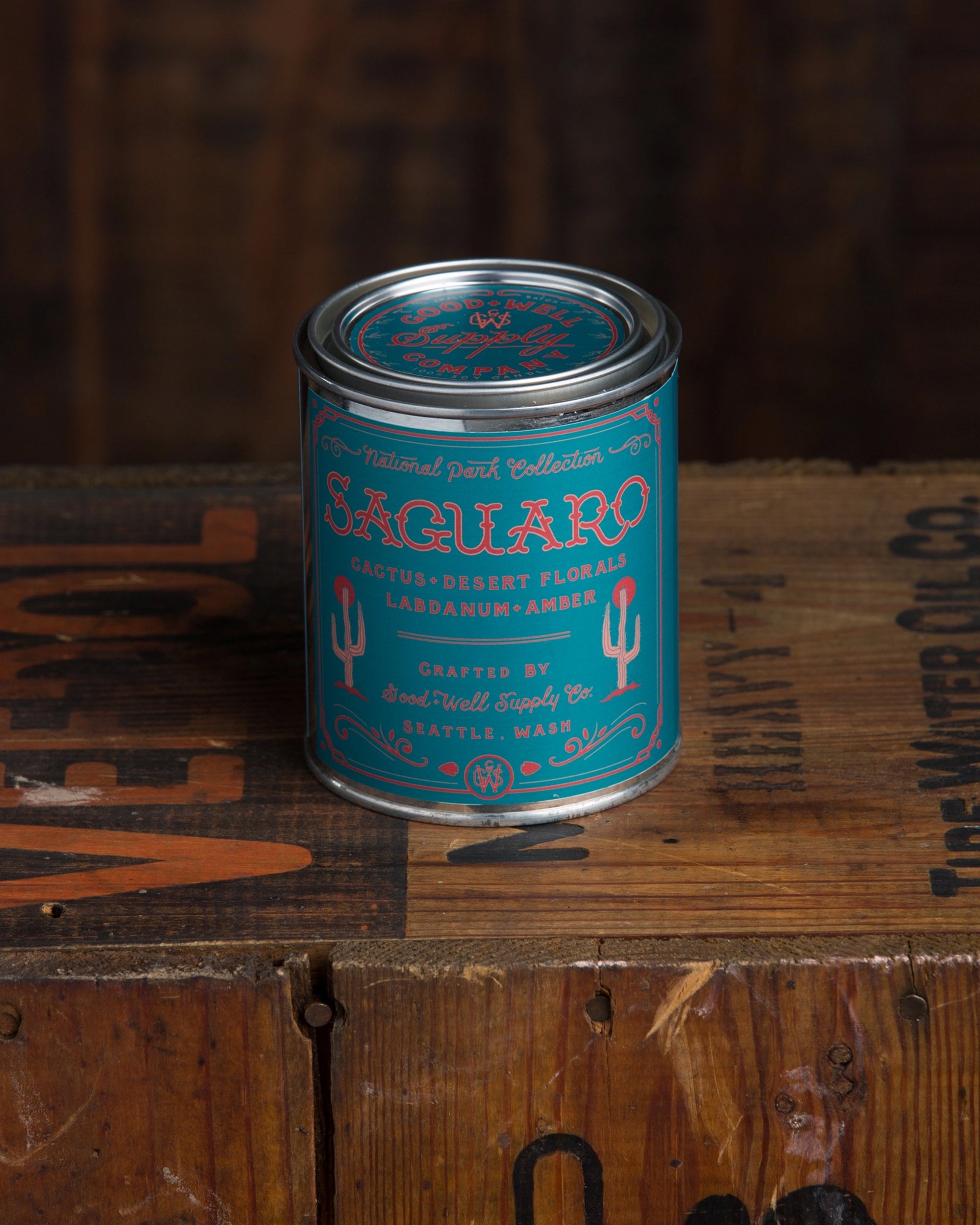 A teal and red "Saguaro National Park Candle" by Faire, featuring cacti graphics and amber notes, rests on rustic wood—evoking the warm essence of desert florals.