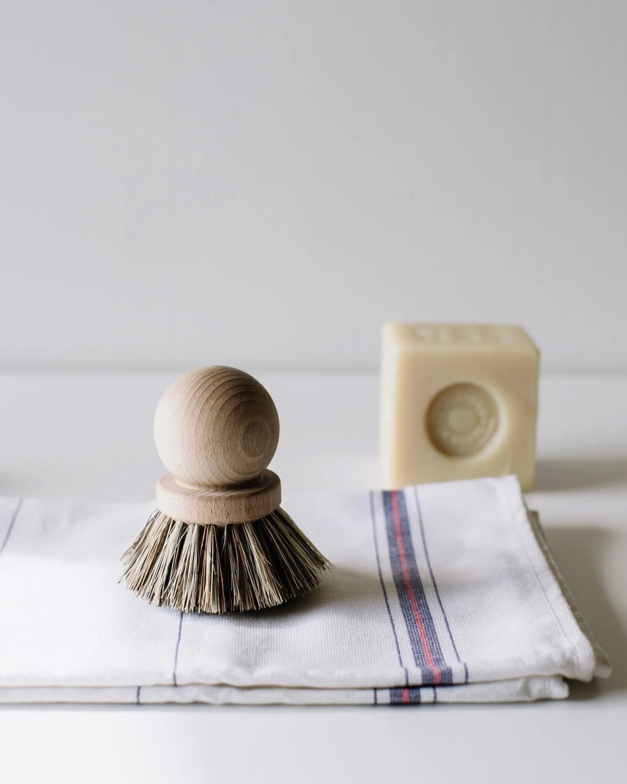 The Be Home 5300-R1819 Saucepan Brush, crafted from French beech wood and natural vegetable fibers, rests on a folded white cloth with red and blue stripes; a rectangular bar of soap appears blurred in the background.