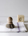 The Be Home 5300-R1819 Saucepan Brush, crafted from French beech wood and natural vegetable fibers, rests on a folded white cloth with red and blue stripes; a rectangular bar of soap appears blurred in the background.