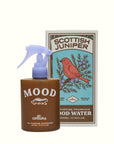 A brown spray bottle labeled "MOOD" by Faire stands before a box with a red bird and juniper berries, displaying "Scottish Juniper Room Spray"—a fabric safe, ethanol-free fragrance for any room.