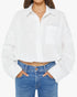 A woman stands against a white background wearing The Scrunchie Frenchie 100% cotton white button-up shirt with a front pocket by Mother, paired with blue jeans and her hands in her jean pockets.