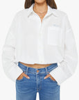 A woman stands against a white background wearing The Scrunchie Frenchie 100% cotton white button-up shirt with a front pocket by Mother, paired with blue jeans and her hands in her jean pockets.