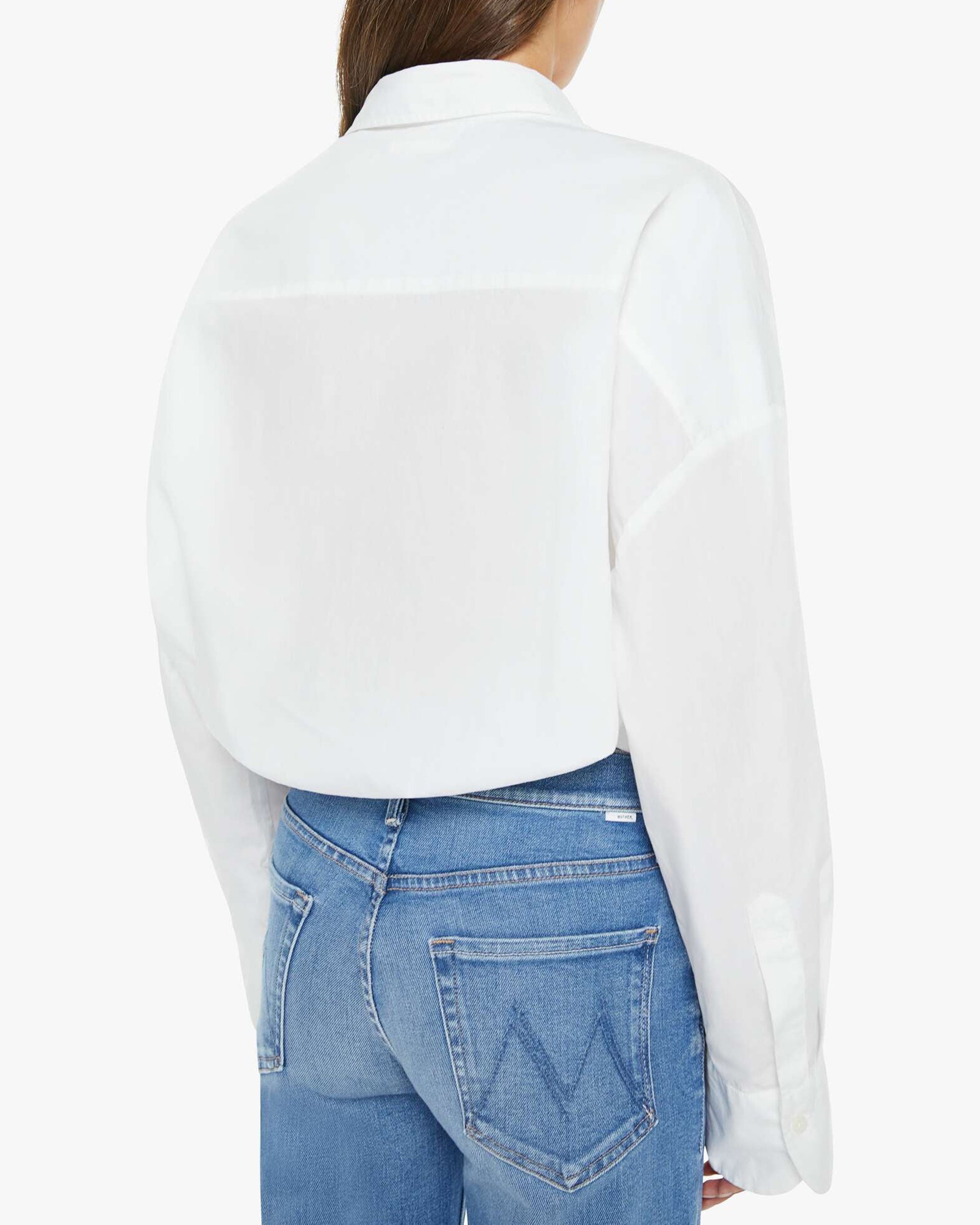 A person is shown from the back wearing Mother’s The Scrunchie Frenchie—an untucked white cotton top with long wide sleeves and light blue jeans featuring “V”-stitched pockets.