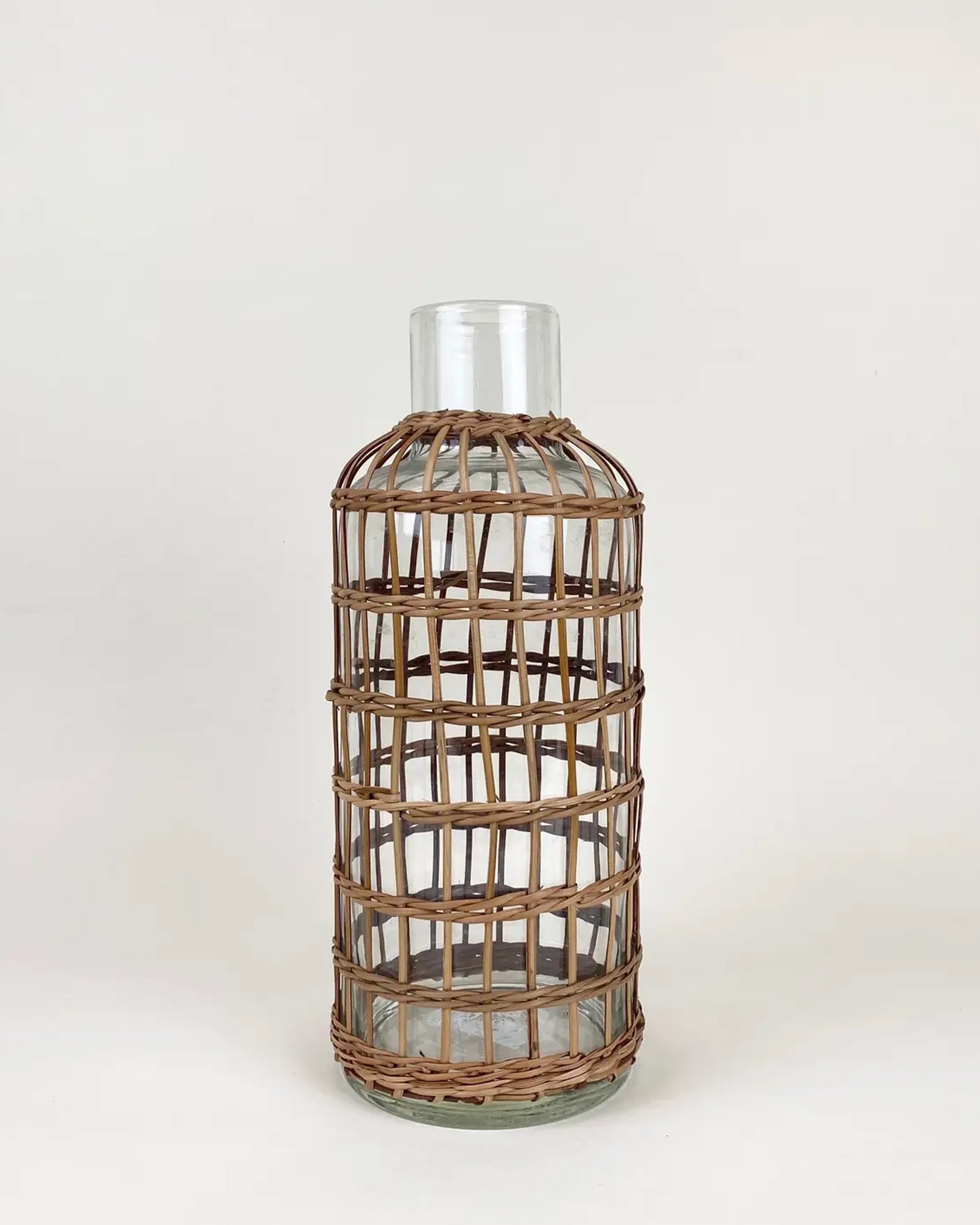 The Be Home Seagrass Rattan Vase M is a clear glass bottle with a handcrafted seagrass rattan sleeve, shown upright against a plain white background.