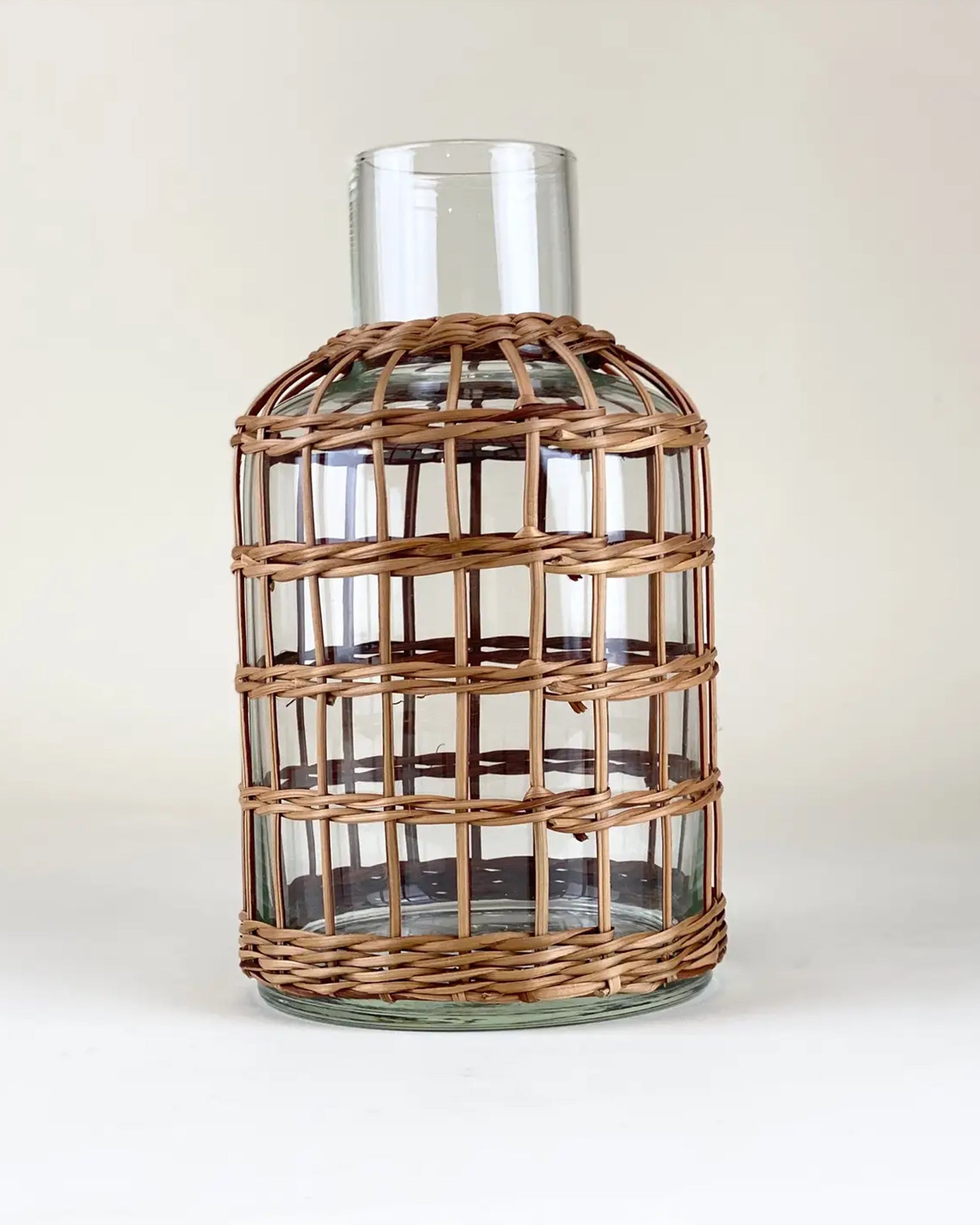 The Be Home Seagrass Rattan Vase S features a clear glass body with a wide neck, wrapped in artisanal seagrass rattan with an open grid pattern—an elegant, handcrafted decor piece ideal for display against an off-white backdrop.