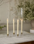 Two cream-colored taper candles from the Serenade Candleholder Set/2 by Accent Decor are displayed in ribbed glass holders on a marble tabletop, with white textured walls and green floral accents in the background.